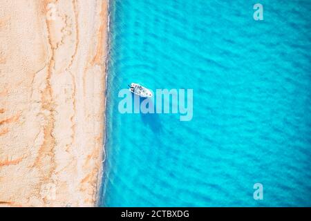 White yacht and turquoise water waves from top view. Beach with yellow sand glowing by sunlight. Travel summer vacations seascape background from drone