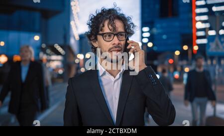 Young Stylish Businessman with Curly Hair is Talking on a Phone While Walking in the City. He's Wearing Glasses. Office People Walk By. It's Evening Stock Photo