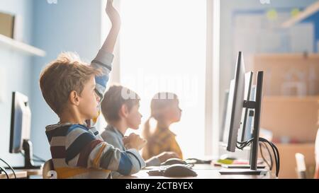 Elementary School Science Classroom: Boy Raises Hand with Question. Teacher Educates Smart Little Schoolchildren who Work on Personal Computers, Learn Stock Photo