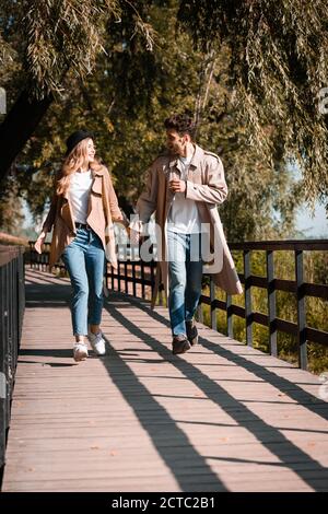 excited couple in trench coats holding hands and walking on wooden bridge Stock Photo