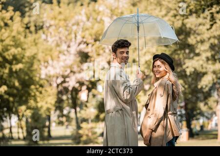 blonde woman in hat and man standing under umbrella in autumnal park Stock Photo