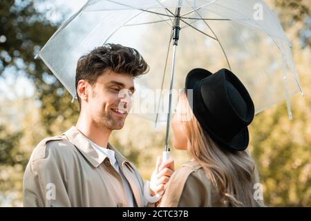 man standing under umbrella and looking at blonde woman in hat in autumnal park Stock Photo