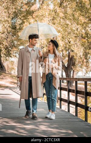 couple in trench coats walking on wooden bridge under umbrella with coffee to go Stock Photo