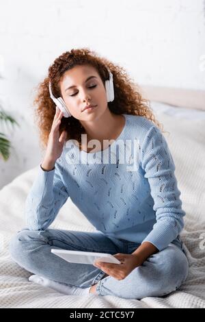 Red haired woman with closed eyes listening music in headphones and holding digital tablet on bed Stock Photo
