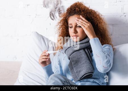 Selective focus of sick woman in scarf holding thermometer on bed Stock Photo
