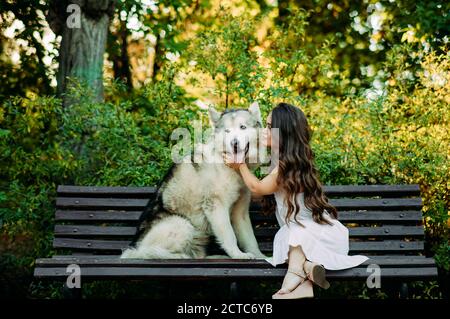 Young woman with innate disorder dwarfism sits on bench next to Malamute dog and hugs it while walking in park. Stock Photo
