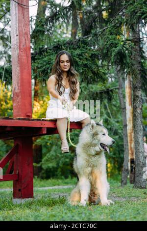 Young woman with innate disorder dwarfism sits on bench next to Malamute dog and keep on a leash it while walking in park. Stock Photo