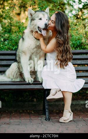 Young woman with innate disorder dwarfism sits on bench next to Malamute dog, hugs and kisses it while walking in park. Stock Photo