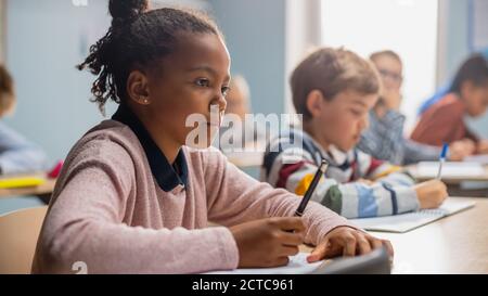 In Elementary School Classroom Brilliant Black Girl Writes in Exercise Notebook, Taking Test and Writing Exam. Junior Classroom with Diverse Group of Stock Photo