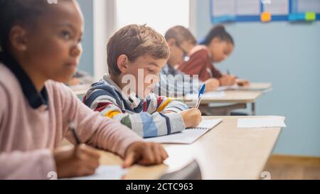 In Elementary School Classroom Brilliant Black Girl Writes in Exercise Notebook, Taking Test and Writing Exam. Junior Classroom with Diverse Group of Stock Photo