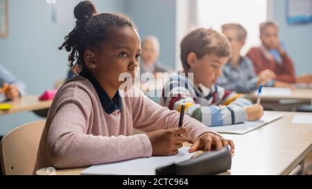 In Elementary School Classroom Brilliant Black Girl Writes in Exercise Notebook, Taking Test and Writing Exam. Junior Classroom with Diverse Group of Stock Photo