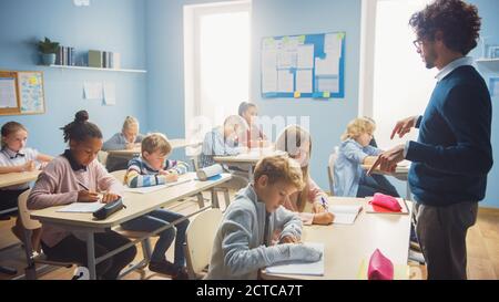 In Elementary School Class: Enthusiastic Teacher walks between Rows of Bright Diverse Children, Dictates Lesson. Group of Smart Multiethnic Kids Stock Photo