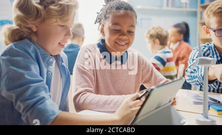 In the Elementary School: Girl and a Boy Work as a Team Using Tablet Computer. Diverse Classroom with Kids Learning Programming Language and Software Stock Photo