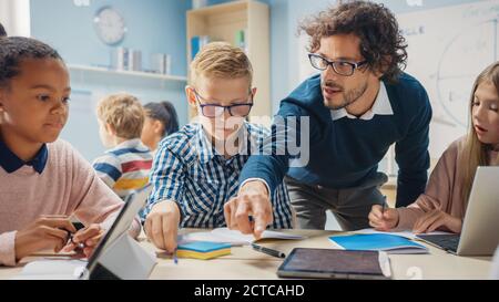 Elementary School Classroom: Enthusiastic Teacher Using Tablet Computer Explains Lesson to Brilliant Young Children. Kids Learning Programming Stock Photo