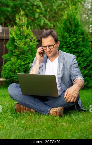 Middle aged businessman sits on grass works on laptop, talks on mobile phone Stock Photo