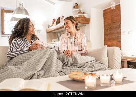 Two fine young women sitting on the sofa with a blanket on their legs laughing enjoying the house in winter having tea with pastries - Mixed race fema Stock Photo