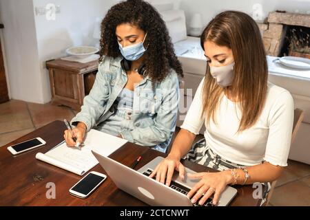 Lifestyle concept, work from home to reinvent your life: two young women, hispanic and caucasian, sitting on a table using a laptop with medical mask Stock Photo
