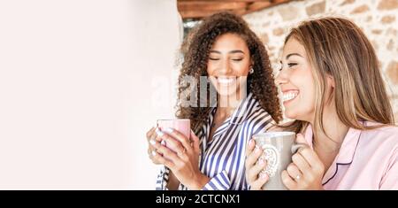Multi-ethnic best friends women in pajama smiling holding a cup of tea with white copy space to the left - Concept: stay at home and enjoy your life Stock Photo