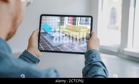 Decorating Apartment: Man Holding Digital Tablet with AR Interior Design Software Chooses 3D Furniture for Home. Man is Choosing Sofa, Table for Stock Photo
