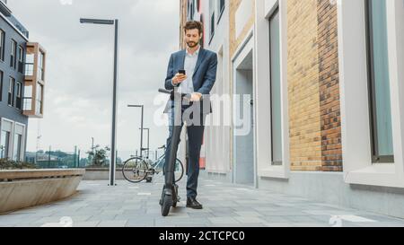 Young Businessman is Checking his Smartphone While RIde to Work on an Electric Scooter. Modern Entrepreneur Uses Contemporary Ecological Transport to Stock Photo