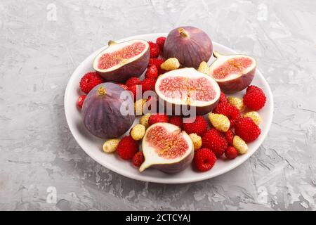 Fresh figs, strawberries and raspberries on white ceramic plate on gray concrete background. side view, close up. Stock Photo