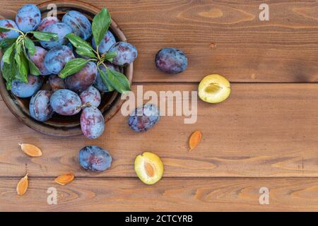 A few washed ripe blue plums with leaves in a brown plate on the wooden table. Several seeds and halves of fruit lie separately. Copy space. The view Stock Photo