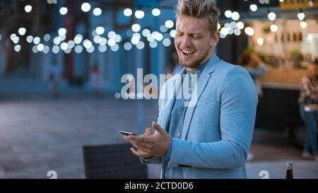 Handsome Happy Young Man is Using a Smartphone while Standing Outdoors in the Evening. He's Browsing the Internet or Social Media, Posting a Status Stock Photo
