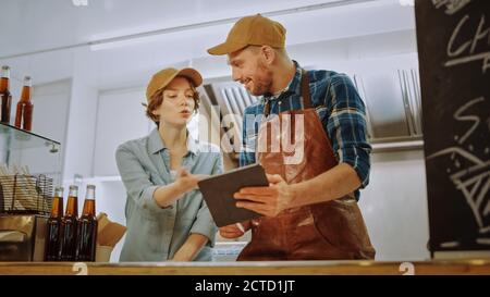 Successful Food Truck Man and Woman Employees in Brown Caps are Using a Tablet Computer. They Discuss Their Profits and Future Business Plans Stock Photo