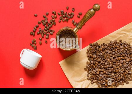 Top view of fresh coffe prepared in  cezve (traditional turkish coffee pot), whte cup and coffee beans on craft paper on red background. Stock Photo