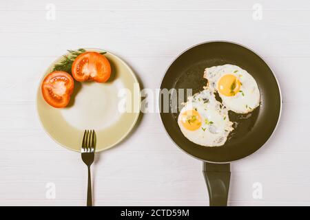 Fried eggs in frying pan next to tomatoes and fork on plate, top view. Stock Photo