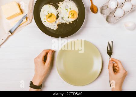 Female hands holding fork next to empty plate among cooking ingredients and frying pan with fried eggs on white table, top view. Stock Photo