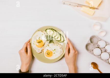 Female hands holding plate fried eggs and fresh cucumber slices next to ingredients on white kitchen table, top view. Stock Photo