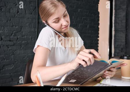 Young woman holds her phone between shoulder and ear and flipping through a book Stock Photo