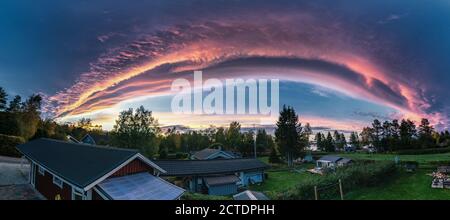 Miraculous panorama of sunset above scandinavian village with rose clouds - edge of cyclone. Arched dark sky with rose border to clear sky. Houses on Stock Photo