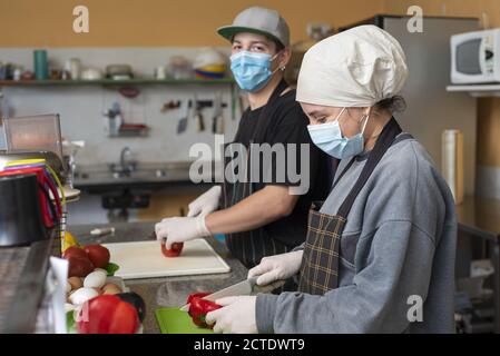 Two young chefs slicing vegetables wearing medical masks Stock Photo