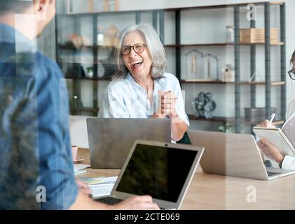 Happy old female mentor talking to young interns at group office meeting. Stock Photo