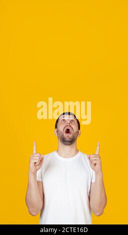 A young man with a beard in a white T-shirt is amazed and surprised, looking up and pointing with his fingers and raised hands. Stands on isolated yellow background. Stock Photo