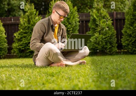 Young hipster guy sits on grass in park working on laptop, checks email Stock Photo