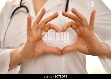 The doctor in a white robe, against the background, shows a gesture with his fingers, in the shape of a heart.  Stock Photo