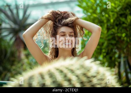 Smiling pretty young woman standing near the cactus and holding her hair Stock Photo