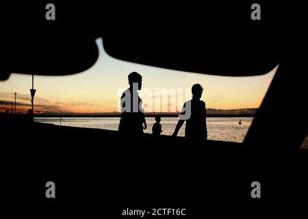 West Cowes seafront at dusk looking through car vehicle windscreen over Solent silhouettes of man woman and child walking along the revetment pavement Stock Photo