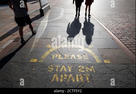 The changing face of the high street. People cast shadows walking past a social distancing information sign painted on the pavement on the high street in Dundee in Scotland, some six months on from the evening of March 23 when Prime Minister Boris Johnson announced nationwide restrictions. Stock Photo