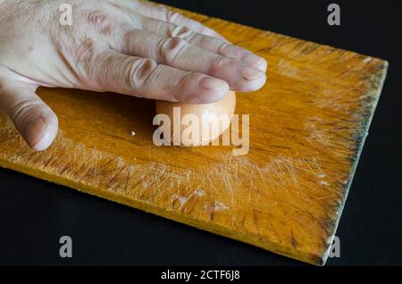 A male hand holds a boiled chicken egg in his shell against the chopping board. Method of cleaning the shells. Cooking. Selective focus. Stock Photo