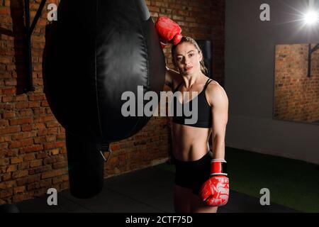 Fit strong brunette woman in sportswear top and shorts fights a punching bag with boxing gloves in the gym Stock Photo