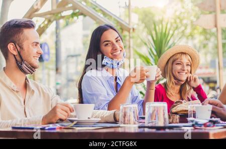 Happy young woman wearing face mask smiling at the camera at the restaurant cafè. Group of multiracial friends having a fun conversation and talking t Stock Photo