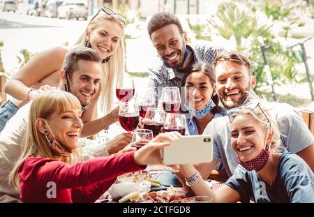 Multiracial group of friends wearing protection mask at the restaurant. Happy people celebrating toasting red wine taking a selfie with smartphone. Stock Photo