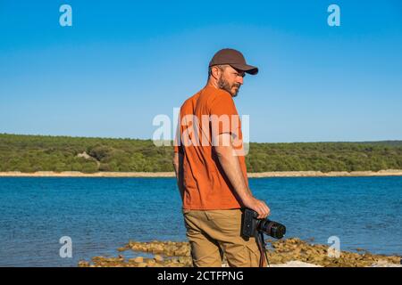 Photographer in t-shirt and cargo pants walking around sea shore with mirror less camera, shooting nature, half body portrait, landscape photography c Stock Photo