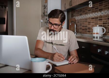 Concentrated millennial hipster guy wearing glasses, listening to favorite music while planning workday. Focused young biracial businessman watching e Stock Photo
