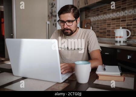 Focused young man wearing glasses using laptop, typing on keyboard, writing email or message, chatting, shopping, successful freelancer working online Stock Photo