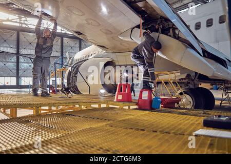 Full length portrait of two engineer workers in uniform fixing and checking commercial airplane in hangar Stock Photo